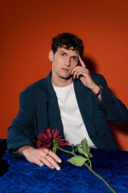 Stylish man touching forehead near burgundy dahlia flower on red background