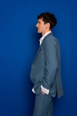 Side view of smiling man in trendy suit posing on blue background 