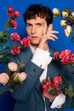 Stylish man in shirt and jacket posing near roses on blue background 