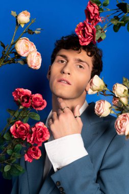 Trendy young man in jacket posing near roses on blue background 