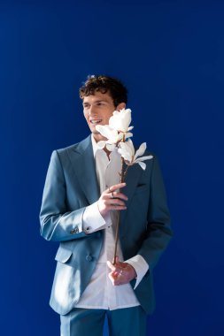 Cheerful curly man holding magnolia flowers isolated on navy blue 
