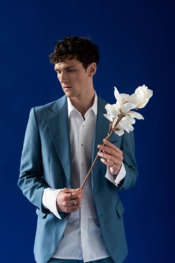 Brunette man in stylish jacket and shirt holding magnolia branch isolated on navy blue 