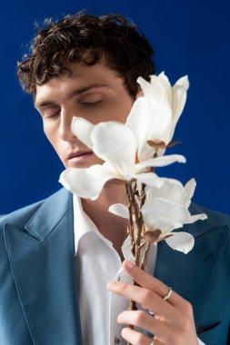 Portrait of curly young man in jacket holding blurred magnolia branch isolated on blue 