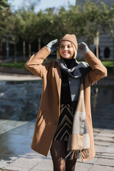 Stock image happy blonde woman in coat and gloves adjusting hat standing smiling outside 