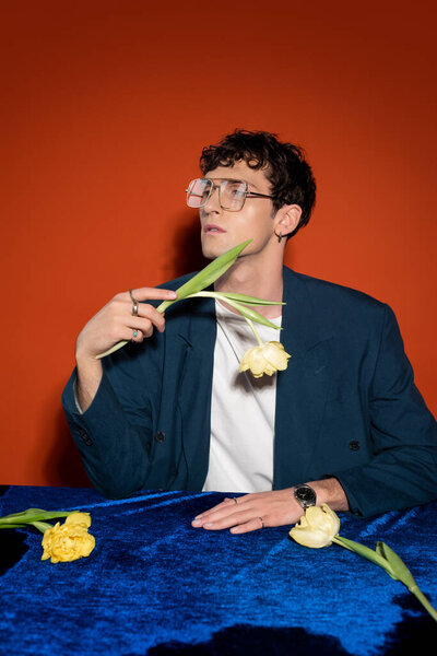 Curly man in blazer and eyeglasses holding tulip on red background