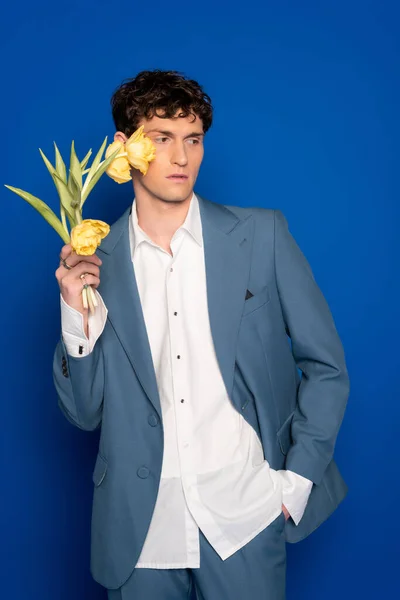 Stylish young man in suit and shirt holding yellow tulips on blue background