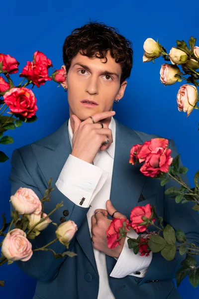 Curly man in stylish outfit touching chin near blooming flowers on blue background