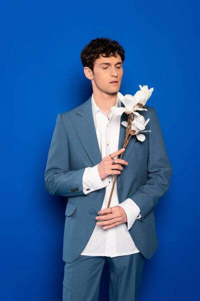 Trendy young man holding magnolia flowers on blue background 