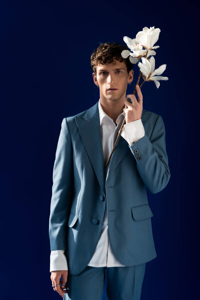 Trendy young man in suit holding branch of magnolia isolated on navy blue 