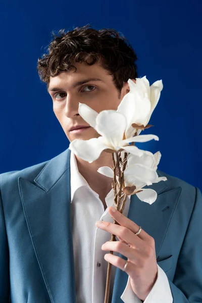 Trendy young man in jacket and shirt holding magnolia flowers isolated on blue