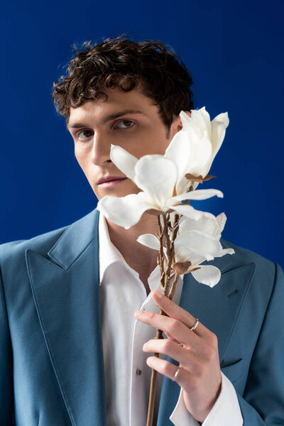 Модный молодой человек в куртке и рубашке держит цветы магнолии изолированы на синий 