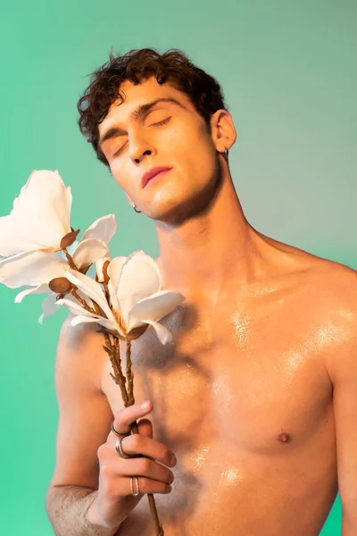Shirtless man with oil on skin holding magnolia flowers on green background