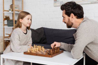Evde sakallı babayla satranç oynarken gülümseyen küçük kız.