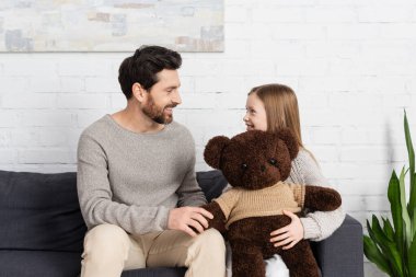 happy girl holding soft toy and looking at cheerful bearded dad while sitting on sofa at home clipart