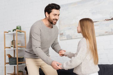 smiling man holding hands of daughter while teaching her to dance at home