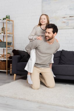 cheerful bearded man piggybacking happy daughter near couch in modern living room