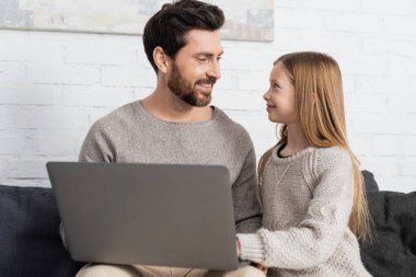 happy father and daughter looking at each other near laptop at home