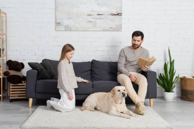 bearded man with book sitting on couch near happy daughter and labrador dog lying on carpet