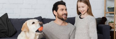 Labrador dog playing with ball near happy father and daughter in living room, banner