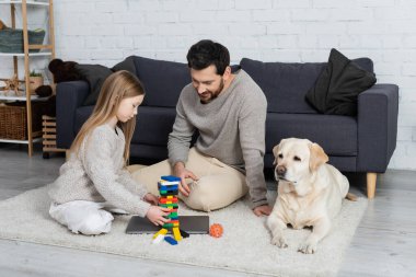 happy bearded man with preteen daughter playing wood blocks game near laptop and labrador dog on floor at home