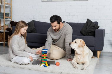 Gülümseyen baba ve kız oturma odasında Labrador köpeğinin yanında tahta blok oyunu oynuyorlar.