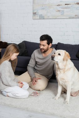 gülümseyen sakallı adamla genç bir kız halının üzerinde oturuyor. Göz rengi palet ve labrador köpeği.