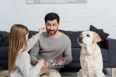 Genç kız elinde makyaj paleti tutarken gülümseyen babasıyla oturma odasında labrador köpeğinin yanında oynuyordu.