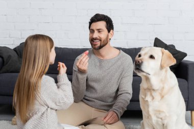 Gülümseyen sakallı adam evde kızıyla ve labrador köpeğiyle oynarken yanında dudak parlatıcısı tutuyor.