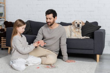 Labrador köpeğinin yanında, oturma odasında kanepede yatarken babasının parmağına yüzük takan bir çocuk.