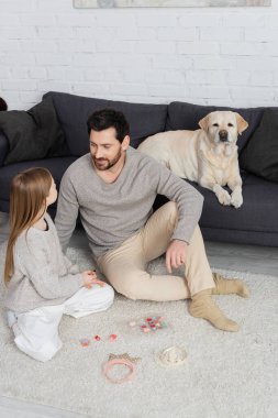 Labrador köpeği babasının ve kızının yanındaki koltukta uzanmış oyuncak taçların ve makyaj paletlerinin yanında yerde oturuyor. 