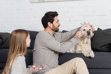 Gülümseyen sakallı adam oyuncak taç takıyor köpeğe, kızının yanında gölge paleti tutuyor.