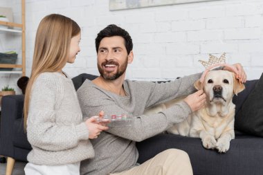 Labrador köpeğine oyuncak taç takan mutlu sakallı bir adam. Yanında küçük kız gölgeler paleti tutuyor.