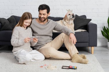 Mutlu baba-kız, oturma odasında Labrador köpeğinin yanında otururken makyaj paletine bakıyor. 