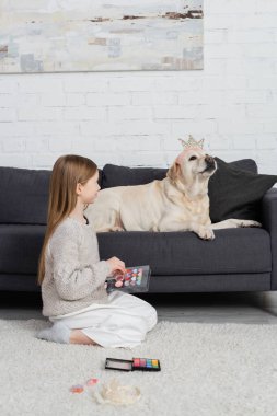 Makyaj paleti tutan mutlu genç kız ve koltukta oturan oyuncak taclı Labrador 'a bakıyor. 