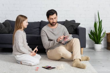 bearded father pointing at makeup palette while sitting on carpet with happy daughter 