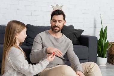 happy girl wearing toy ring on finger of father in crown while playing in living room 