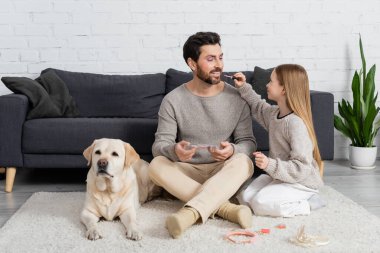 Mutlu kız babasının dudaklarına dudak parlatıcısı sürerken halının üzerinde Labrador 'la oturuyor. 