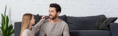 happy girl applying lip gloss on lips of father while sitting near couch, banner 