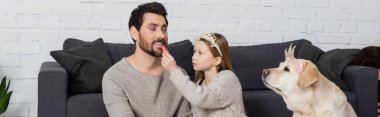 happy girl in toy crown on head sitting near labrador and applying lip gloss on lips of father, banner  clipart