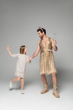 full length of girl in dress holding hands with dad in dress and toy wand on grey  clipart