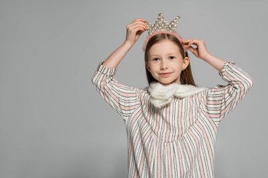 happy girl in dress adjusting crown on head and smiling isolated on grey 