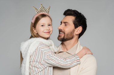 cheerful kid in crown looking at camera while hugging father isolated on grey  clipart