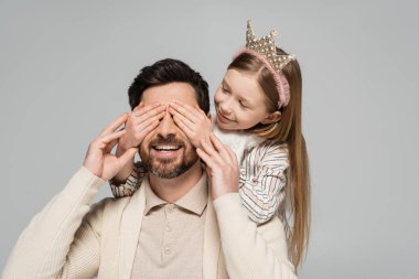cheerful kid in crown covering eyes of bearded father isolated on grey 