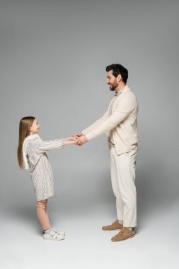 side view of happy father and daughter holding hands while looking at each other on grey  clipart