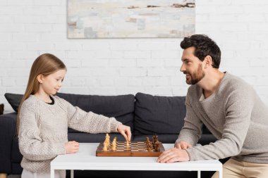 side view of clever girl playing chess with cheerful father in living room 