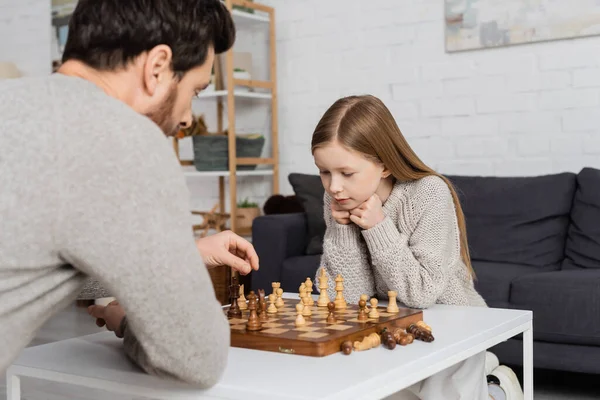 preteen girl thinking near dad and chessboard on coffee table in living room
