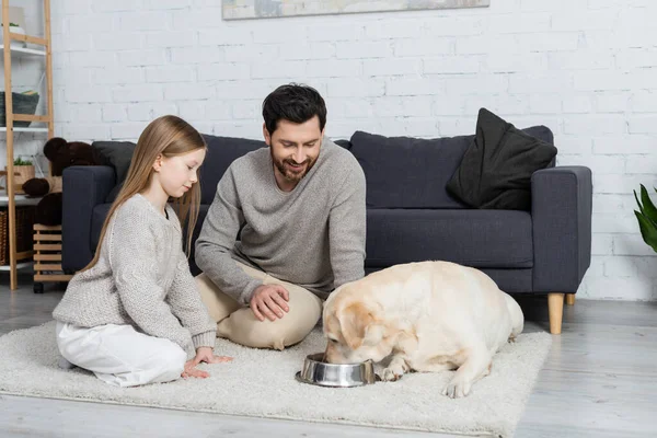 labrador dog drinking water from bowl near cheerful father and daughter on floor carpet at home