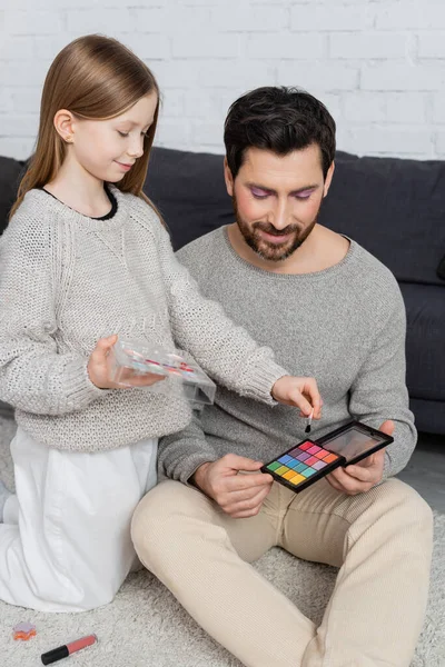 happy father holding eye shadow palette near daughter with cosmetic brush playing beauty game