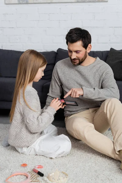 happy father pointing at eye shadow palette near daughter in living room