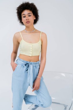 Genç Afrikalı Amerikalı kadın üstte ve pantolonu gri sandalyenin yanında duruyor. 